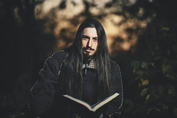 万圣节的时候 一个长头发 留着胡子 穿着黑色斗篷的男人就像一个巫师术士 黑暗魔术师是森林中的一个人 一个巫师在黑暗的森林里带着一本魔法书 — 图库照片