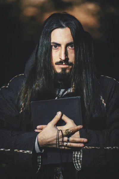 ハロウィーンの魔術師のウォーロックのイメージで黒いマントの長い髪とひげを持つ男 闇の魔術師は 森の中の男 魔法の本を持つ暗い森の中のウィザード — ストック写真