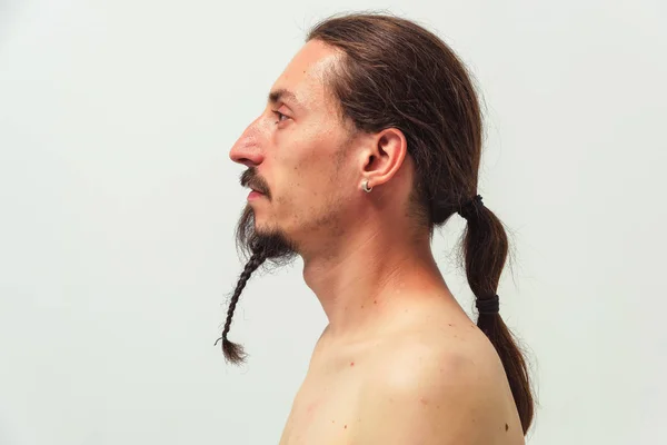 A man with long hair and a beard with a naked torso on white background side view in profile. Long hair gathered in a ponytail and a beard braid at the young man.