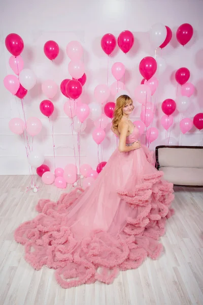 A young woman in a pink ball gown on a background of balloons. Beautiful girl in evening puffy Cloud dress with pink and white balloons. Air lush ball gown on the girl