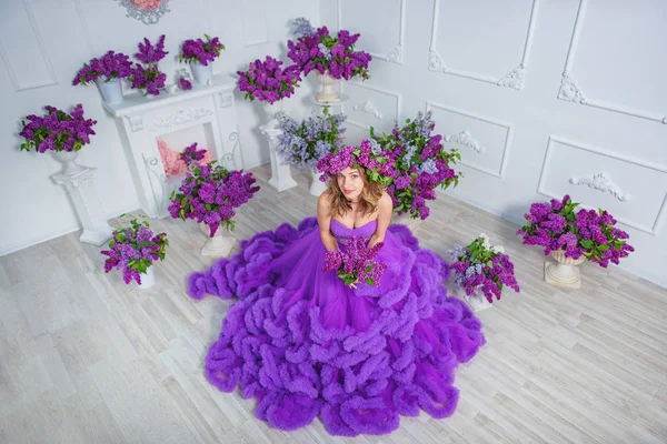 A girl in a lush evening lilac dress on a background of lilacs. A purple cloud dress on a young woman. Beautiful happy smiling girl in a ball gown on a background of lilac flowers.