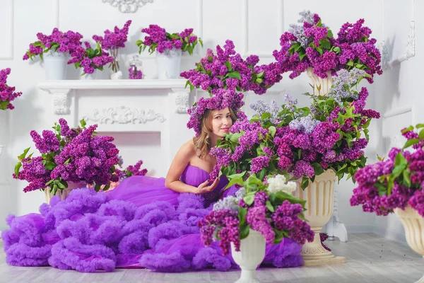 A girl in a lush evening lilac dress on a background of lilacs. A purple cloud dress on a young woman. Beautiful happy smiling girl in a ball gown on a background of lilac flowers.