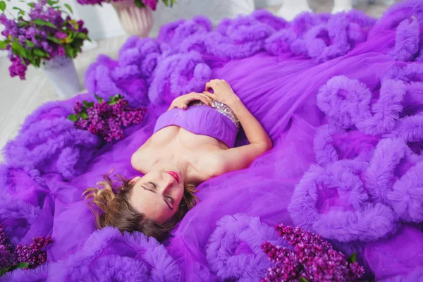 A girl in a lush evening lilac dress lies on the floor. A purple cloud dress on a young woman. Beautiful happy smiling girl in ball gown.