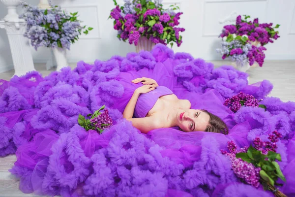 A girl in a lush evening lilac dress lies on the floor. A purple cloud dress on a young woman. Beautiful happy smiling girl in ball gown.