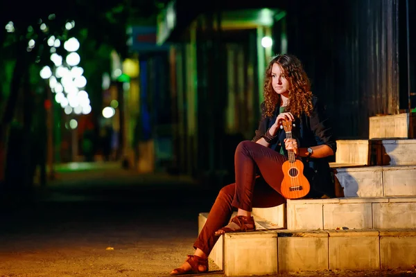 Girl with ukulele in the night city. Young woman with small guitar on walk in night city. A girl with a musical instrument ukulele performs on the streets of the city