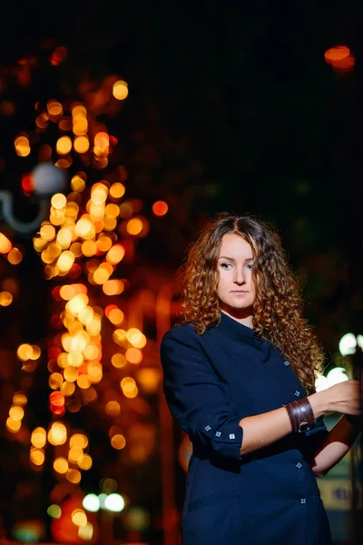 Portrait of a young beautiful woman against the night lights of the city. Red-haired girl on the background of fiery red yellow bokeh. Fabulous portrait of a girl on the background of Christmas lights.