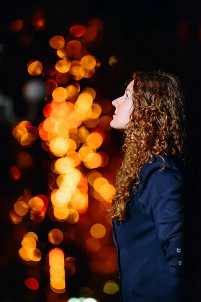 Portrait of a young beautiful woman against the night lights of the city. Red-haired girl on the background of fiery red yellow bokeh. Fabulous portrait of a girl on the background of Christmas lights.