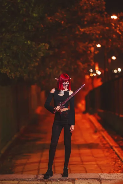 Girl cosplay in the image of a dark elf in a red wig with a sword on the background of the night city. A woman with red hair in an elf costume with false ears and makeup. Dark elf outfit for Halloween.