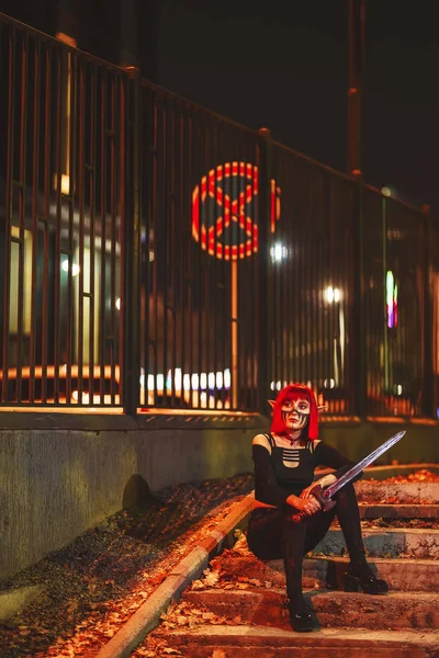 Girl cosplay in the image of a dark elf in a red wig with a sword on the background of the night city. A woman with red hair in an elf costume with false ears and makeup. Dark elf outfit for Halloween.