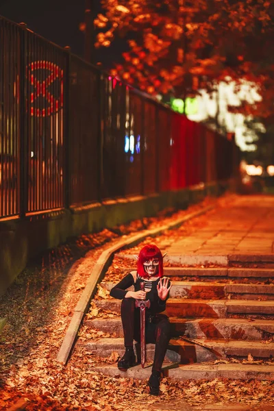 Girl cosplay in the image of a dark elf in a red wig with a sword on the background of the night city. A woman with red hair in an elf costume with false ears and makeup. Dark elf outfit for Halloween.