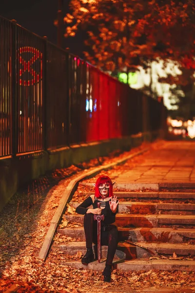 Girl cosplay in the image of a dark elf in a red wig with a sword on the background of the night city. A woman with red hair in an elf costume with false ears and makeup. Dark elf outfit for Halloween.