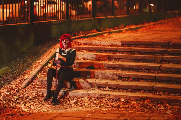 Girl cosplay in the image of a dark elf in a red wig with a sword on the background of the night city. A woman with red hair in an elf costume with false ears and makeup. Dark elf outfit for Halloween.