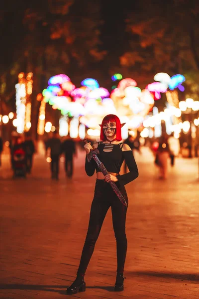 Girl cosplay in the image of a dark elf in a red wig with a sword on the background of the night city. A woman with red hair in an elf costume with false ears and makeup. Dark elf outfit for Halloween.
