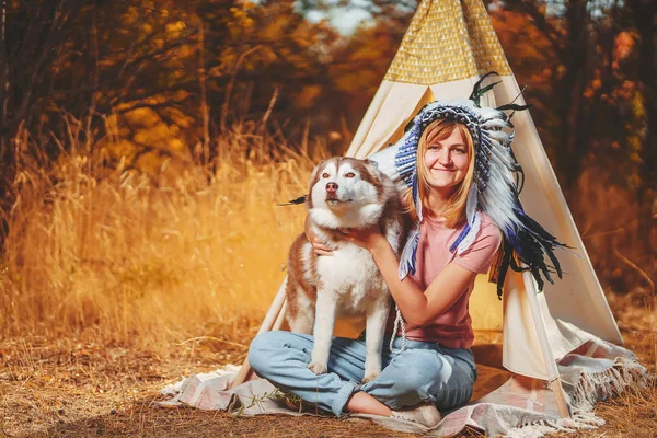 A girl with an Native American crown of feathers on her head with a dog on the background of children\'s wigwam. A young woman in an Indian costume with red husky. Girl with Feather Headdress themed photo shoot
