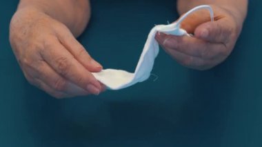 Yaşlı bir kadının ellerinde gazlı bez maskesi. Tıbbi maske takan yaşlı bir kadının elleri. Gazlı bezden yapılmış ev yapımı tıbbi maske.