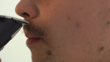 Genç bir adam sakalını elektrikli jiletle tıraş ediyor. Adam beyaz arka planda tıraş oluyor. Yüzünde saç kesimi var..
