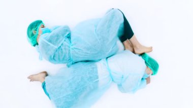 Koruyucu takım elbiseli ve yüz maskeli iki kadın. Genç kızlar koruyucu sağlık önlükleri, kepleri ve maskeleriyle beyaz zeminde yatıyorlar. Grip salgını trajedisi