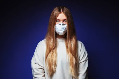 Tıbbi koruyucu maske takmış uzun saçlı güzel bir kız. Mavi arka planda maskeli genç bir kadın. Salgın virüsüne karşı solunum cihazındaki kız