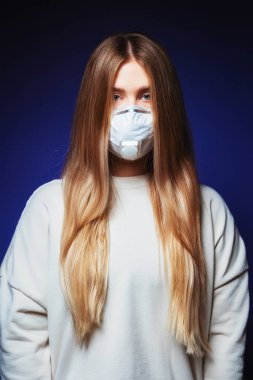 Tıbbi koruyucu maske takmış uzun saçlı güzel bir kız. Mavi arka planda maskeli genç bir kadın. Salgın virüsüne karşı solunum cihazındaki kız