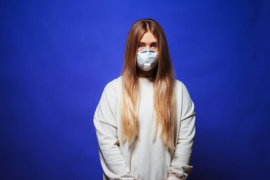 Tıbbi koruyucu maske takmış uzun saçlı güzel bir kız. Mavi arka planda maskeli genç bir kadın. Salgın virüsüne karşı solunum cihazındaki kız