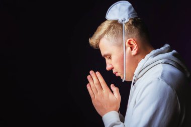 Kafasında tıbbi koruyucu maske olan genç bir adamın portresi. Salgını sona erdirmek için dua et. Anlamsız tıbbi maske kullanımı