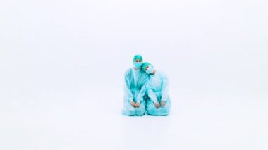 Koruyucu takım elbiseli ve yüz maskeli iki kadın. Genç kızlar koruyucu sağlık önlükleri, kepleri ve maskeleriyle beyaz bir zeminde diz çöküyorlar. Grip salgını trajedisi