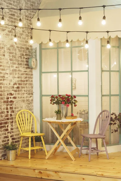 Summer porch with garlands of light bulbs. Terrace of the summer house design of the photo Studio. A verandah patio with flowers roses in metal buckets and wooden chairs and a table.