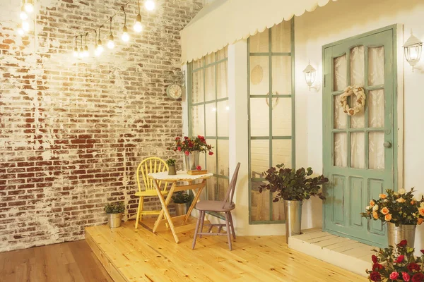 Summer porch with garlands of light bulbs. Terrace of summer house design of photo Studio. Patio verandah with flowers in metal buckets and wooden chairs and table. Street garland with light bulb