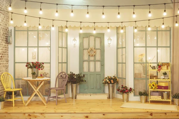 Summer porch with garlands of light bulbs. Terrace of summer house design of photo Studio. Patio verandah with flowers in metal buckets and wooden chairs and table. Street garland with light bulb