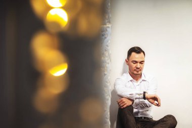 Asyalı adam, Doğu tarzı Bokeh çelenklerinden oluşan beyaz bir duvarın yanında oturuyor. Erkek model güzel bir portre. Resmi olmayan bir ortamda beyaz bir arkaplanda eli kolunda bir iş adamı.