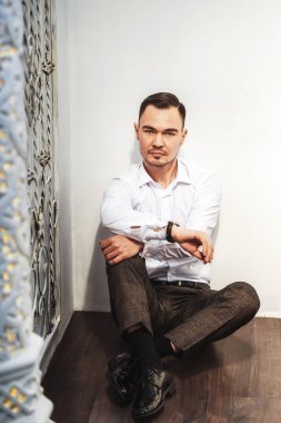  Asyalı bir adam beyaz zeminde oturuyor. Doğu tarzında. Genç adam güzel bir portre modeli. Gömlekli bir işadamı ve beyaz arka planda elinde bir saatle gayri resmi bir ortamda