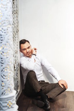  Asyalı bir adam beyaz zeminde oturuyor. Doğu tarzında. Genç adam güzel bir portre modeli. Gömlekli bir işadamı ve beyaz arka planda elinde bir saatle gayri resmi bir ortamda