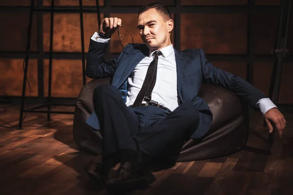 Portrait of a man in a white shirt and suit with glasses in his hands on a dark wooden background. Beautiful stylish male sexy flirting look portrait in a loft style room. Young Asian gentleman