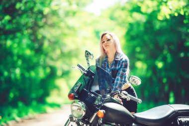 Bahçedeki parkın arka planında motosikletli bir kız. Yeşil ağaçların arkasında bir kadın ve bir motosiklet. Motosikletli şık bir kadın.