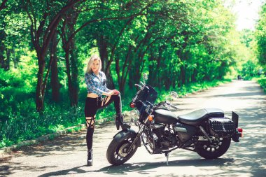 Bahçedeki parkın arka planında motosikletli bir kız. Yeşil ağaçların arkasında bir kadın ve bir motosiklet. Motosikletli şık bir kadın.
