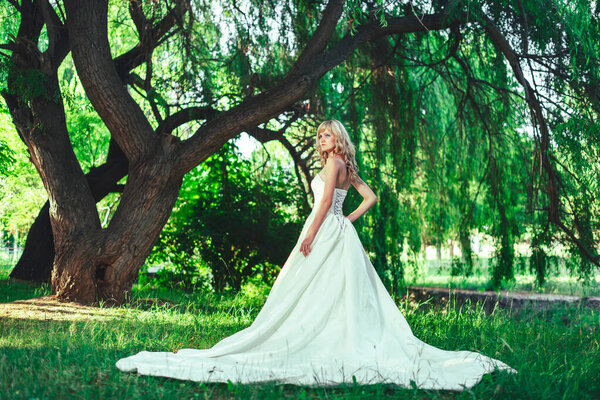 Girl in a wedding dress with a train in the garden Park. A woman in a white evening ball gown in the woods. Bride in a long dress against the background of green trees