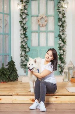 Noel süslemelerinin arka planında köpekli bir kız var. Noel verandasında beyaz köpeği olan bir kadın. Beyaz köpekli Asyalı kadın.