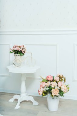 Beyaz bir duvara karşı çiçekli küçük bir sehpa. İç kısımda bisiklet şeklinde saksıda duran pembe çiçekler var. Beyaz vazodaki yapay pembe sahte çiçekler.