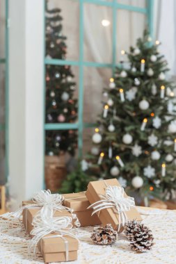 Noel ağacının arka planında karla kaplı kozalaklı kutular sunuluyor. Verandada beyaz kurdeleli açık kahverengi karton kutular ve Noel süslerinin arka planında fiyonklar