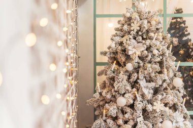 Tasarımcı beyaz Noel ağacı ve ön planda Bokeh çelenkleri. Karla kaplı Noel ağacı, beyaz süsler ve büyük bir pencerenin arkasında gümüş noktalar. Güzel bir Noel kartı geçmişi