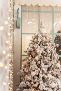 Tasarımcı beyaz Noel ağacı ve ön planda Bokeh çelenkleri. Karla kaplı Noel ağacı, beyaz süsler ve büyük bir pencerenin arkasında gümüş noktalar. Güzel bir Noel kartı geçmişi
