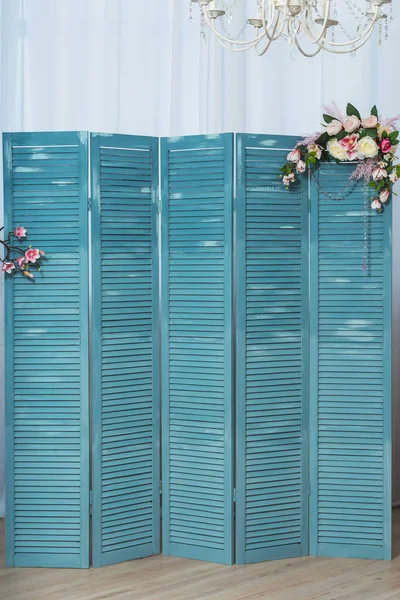 The texture of the slats of a blue wooden folding screen with glass beads and floral arrangement decor. Casement Shutters close-up. Wooden louvre. Interior louver folding screen with flowers