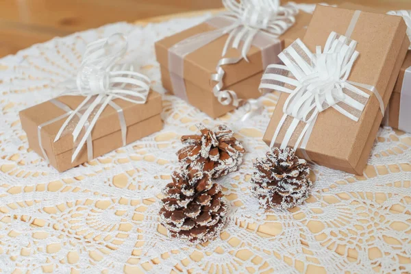 Square craft gifts boxes with snow-covered cones on the table. Light brown cardboard boxes with white ribbons and bows Christmas gifts. New year gifts on a white knitted tablecloth