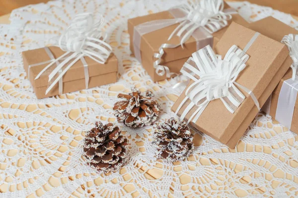 Square craft gifts boxes with snow-covered cones on the table. Light brown cardboard boxes with white ribbons and bows Christmas gifts. New year gifts on a white knitted tablecloth