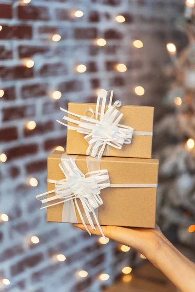 Gift craft boxes on the palm of your hand close-up against a brick wall. Gift box with a white bow and ribbons on the background of bokeh warm garlands. Christmas gift on a woman\'s hand