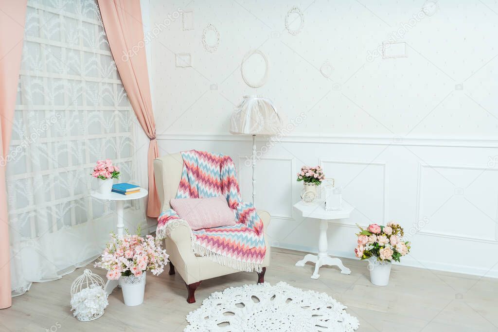 A small cozy living room in the style of shabby chic with a beige armchair. An armchair with a pink blanket and a floor lamp in the room is a classic interior with a spring decor. Frames on the wall. Flowers in white pots vases