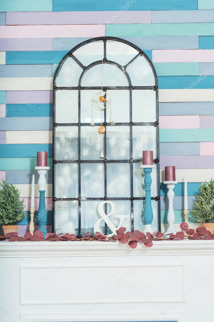Wooden painted candlesticks on a fireplace with a mirror. Spring decoration in the living room. Mirror in the form of a french window in the interior against the backdrop of a multi-colored wall