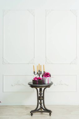 Siyah bir masanın üzerindeki şamdanda altın mumlar, beyaz bir duvarın arka planında alçı pervazlı hediye kutuları. Kahve masasında kurdeleli pembe kurdeleli siyah ve gümüş ambalaj kağıtları..