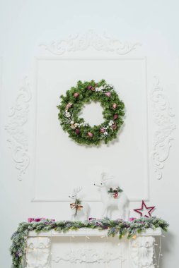 Şöminedeki iki beyaz noel geyiği figürü. Noel geyiği ve köknar dallarıyla şömine dekorasyonu. Şöminenin üzerinde pembe koni olan Noel çelengi.