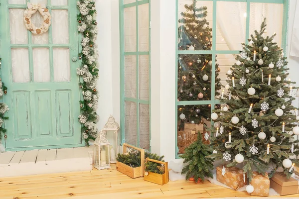 Porch with green door in Christmas decorations and Christmas trees. Spruce garlands around the door. Beautiful winter terrace of the house with garlands of retro light bulbs